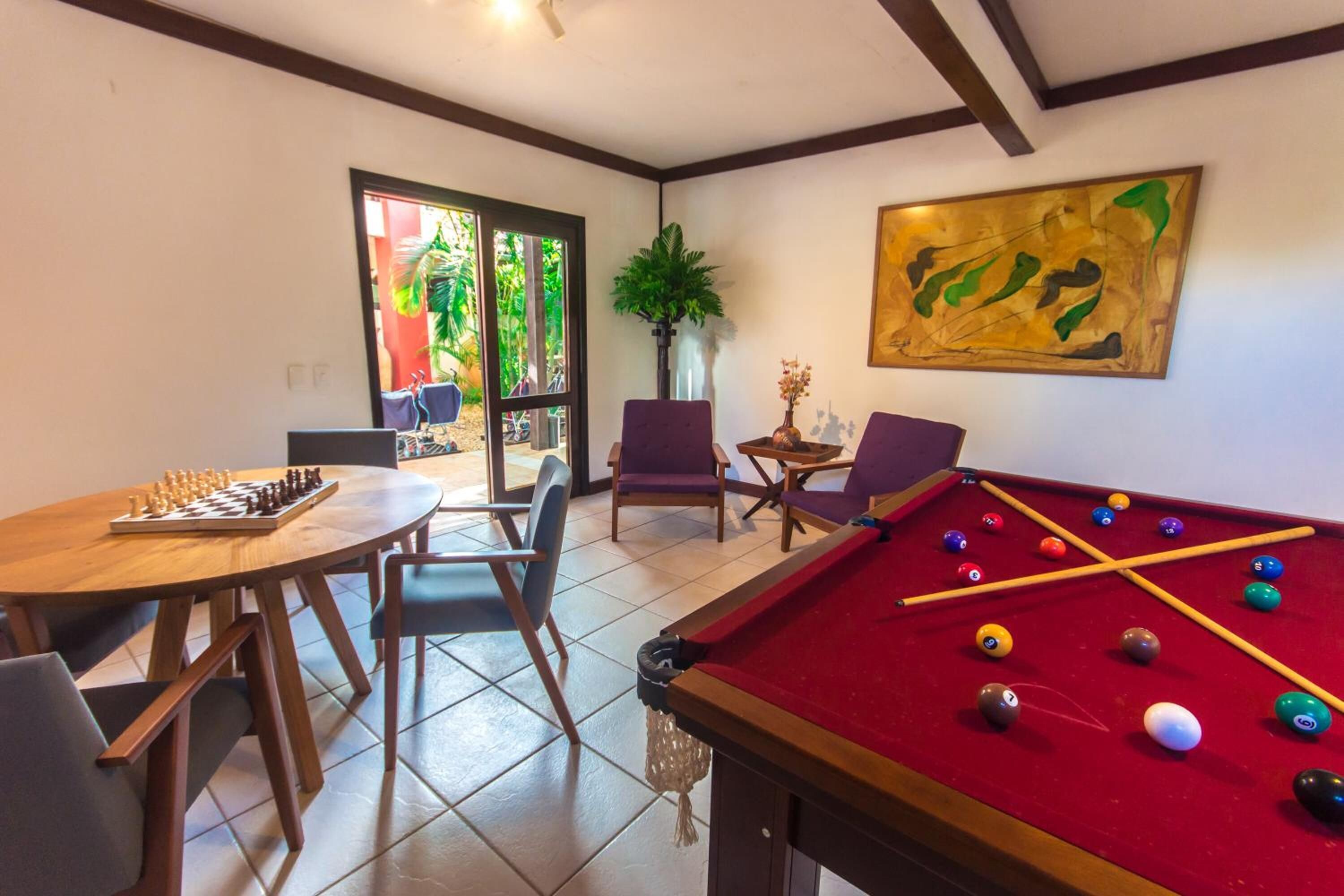 A pool table with billiard balls, a pool cue, and a chessboard on a wooden table. There are chairs around the table, and a potted plant is visible in the background.
