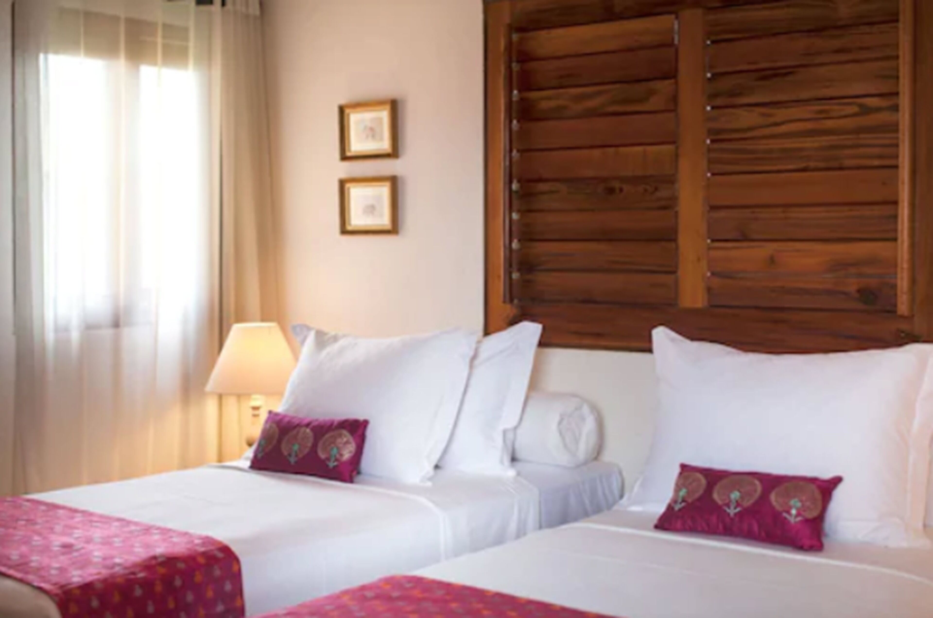 A hotel room with two beds, a wooden headboard, a window with curtains, and two framed pictures on the wall.