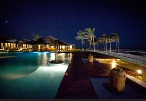 A nighttime resort pool area with overwater bungalows and palm trees.
