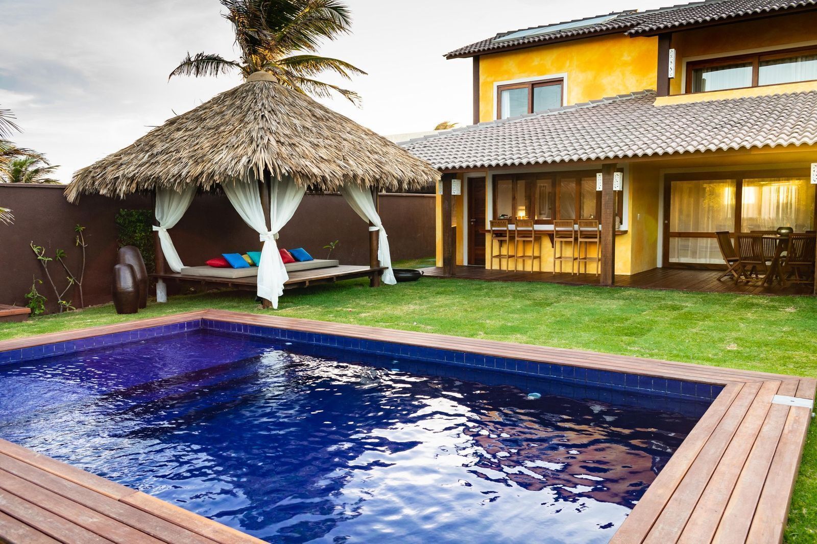 Uma piscina com deck de madeira, área de estar ao ar livre coberta por um telhado de palha e um edifício amarelo com telhado de azulejos.