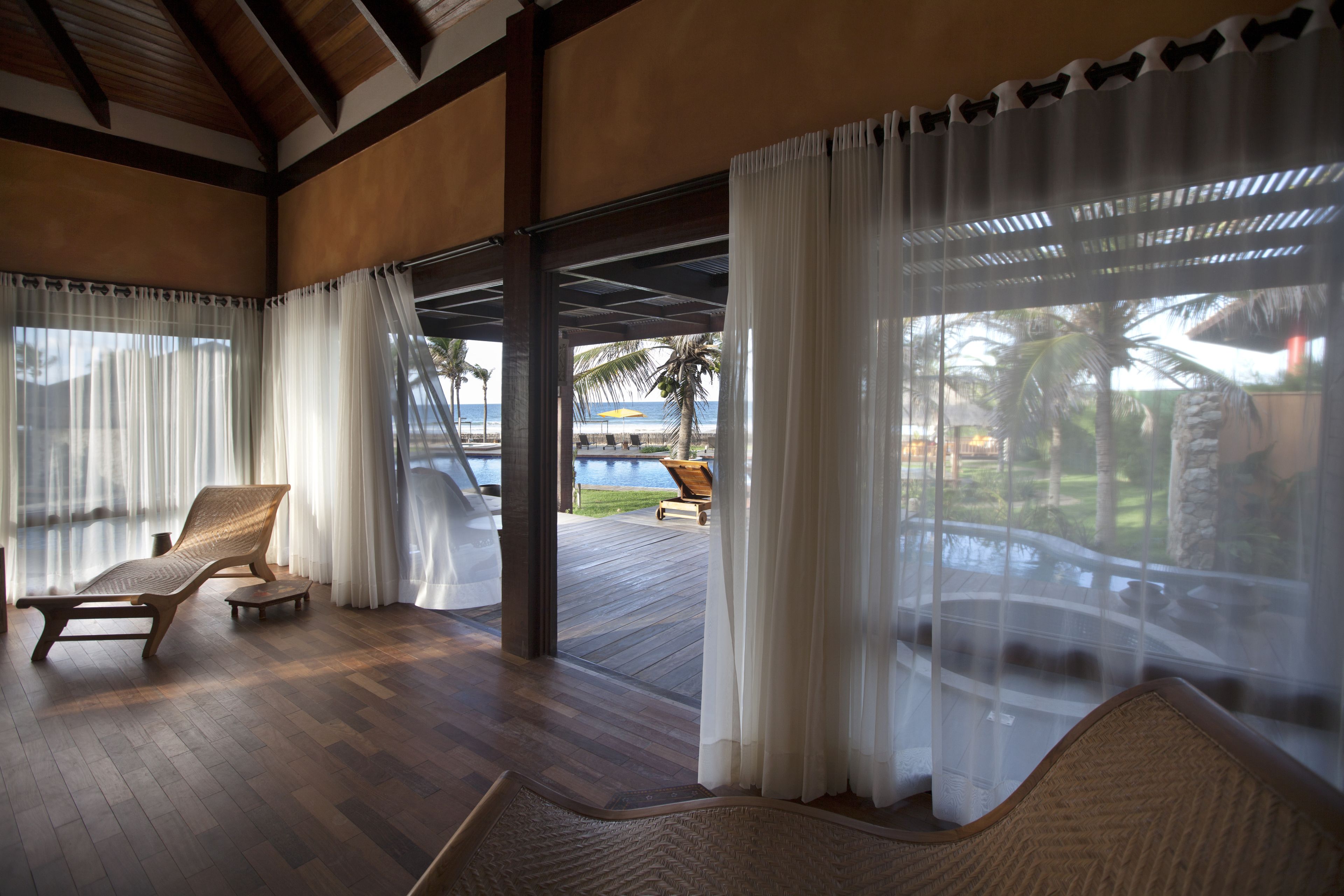 A spacious room with wooden flooring, two lounge chairs, and a view of a pool and palm trees.