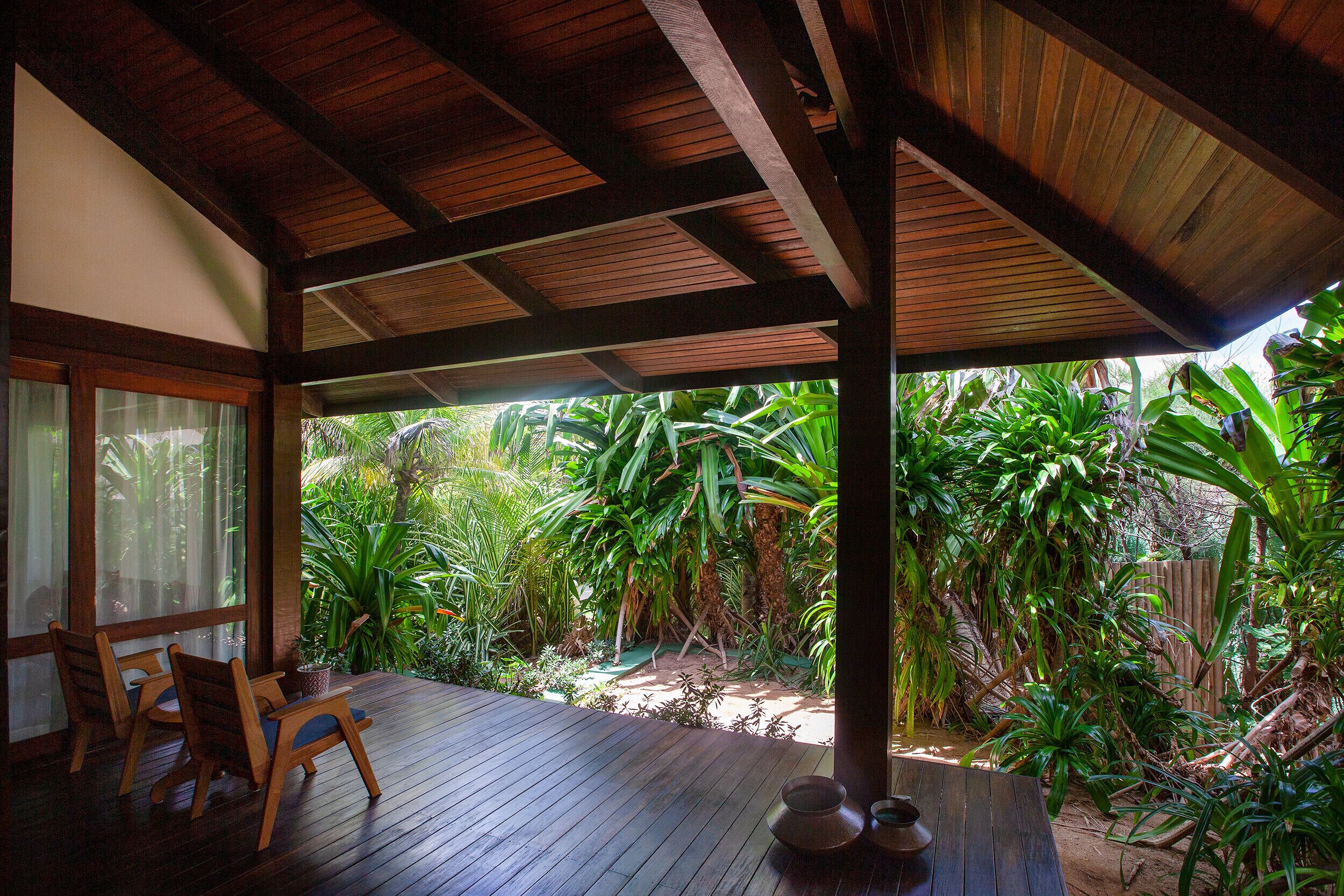A wooden deck with chairs and a table, surrounded by lush tropical plants and a wooden fence.