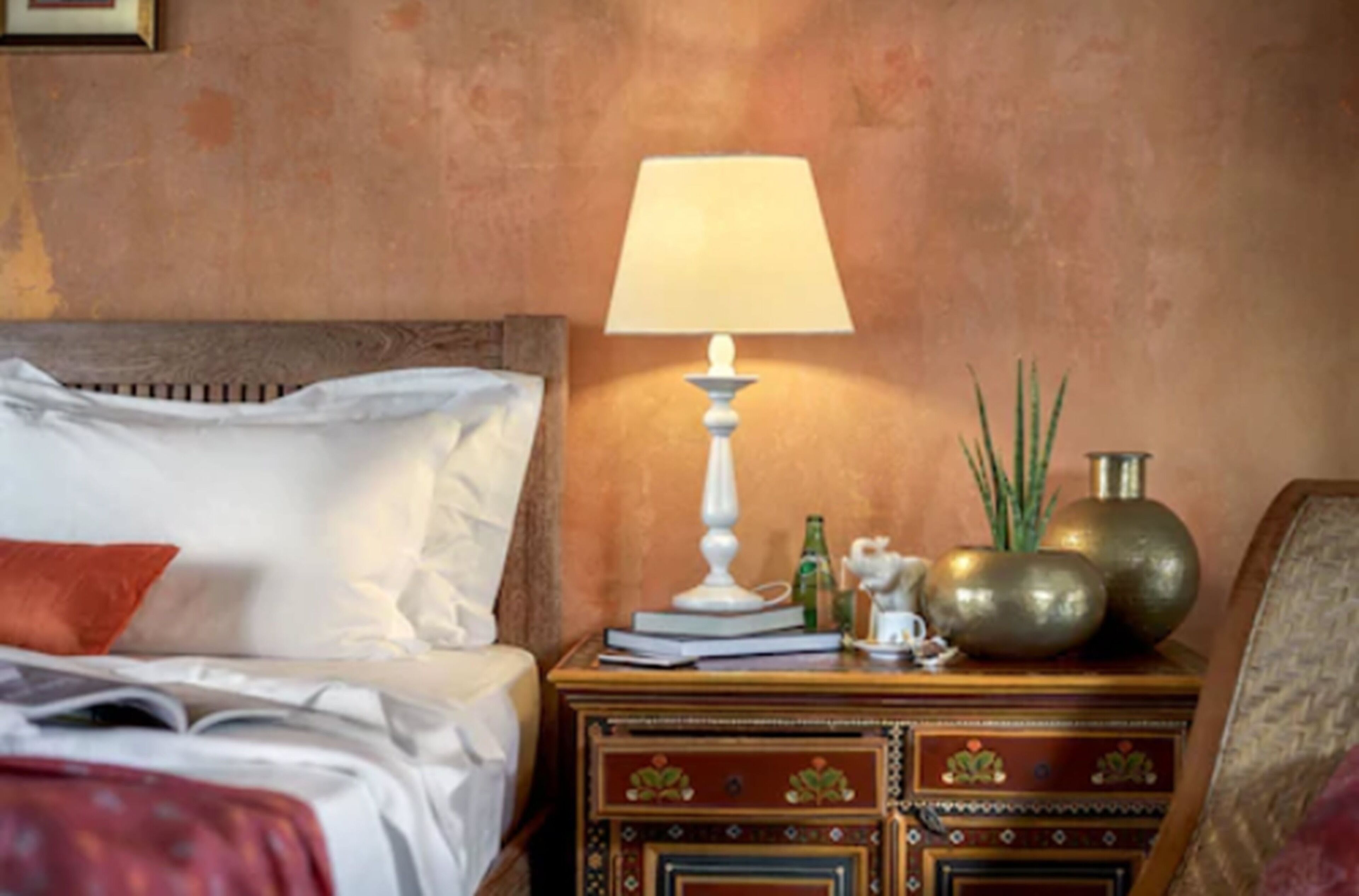 Una cama bien hecha con almohadas blancas y rojas, una mesita de noche con lámpara, libros y objetos decorativos.