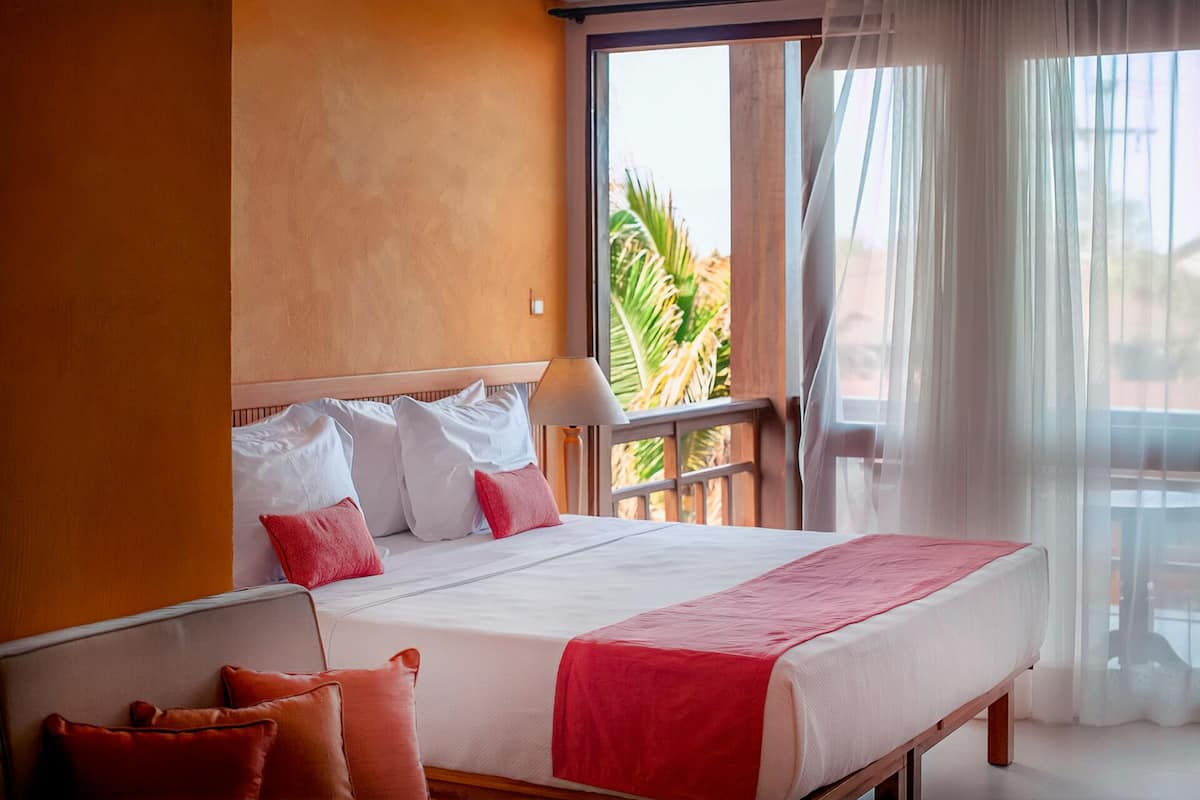 Ein Hotelzimmer mit einem großen Bett, weißer Bettwäsche und einer roten Tagesdecke. Ein Fenster bietet Blick auf Palmen und ein Balkon mit weißem Vorhang.