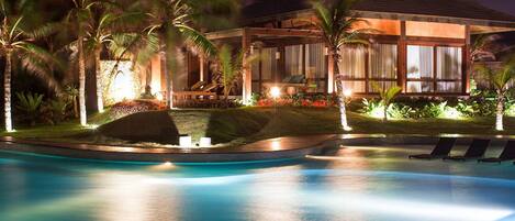 A nighttime view of a resort with a swimming pool, palm trees, and a well-lit building with a roof.