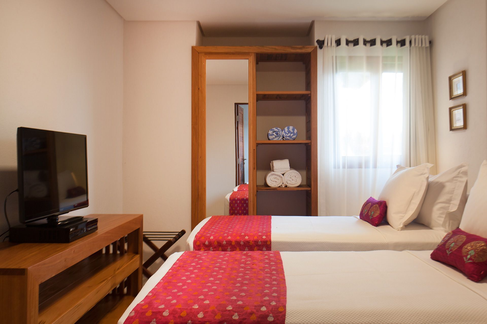 Habitación de hotel con dos camas, televisión, estante de madera, armario y ventana con cortinas.