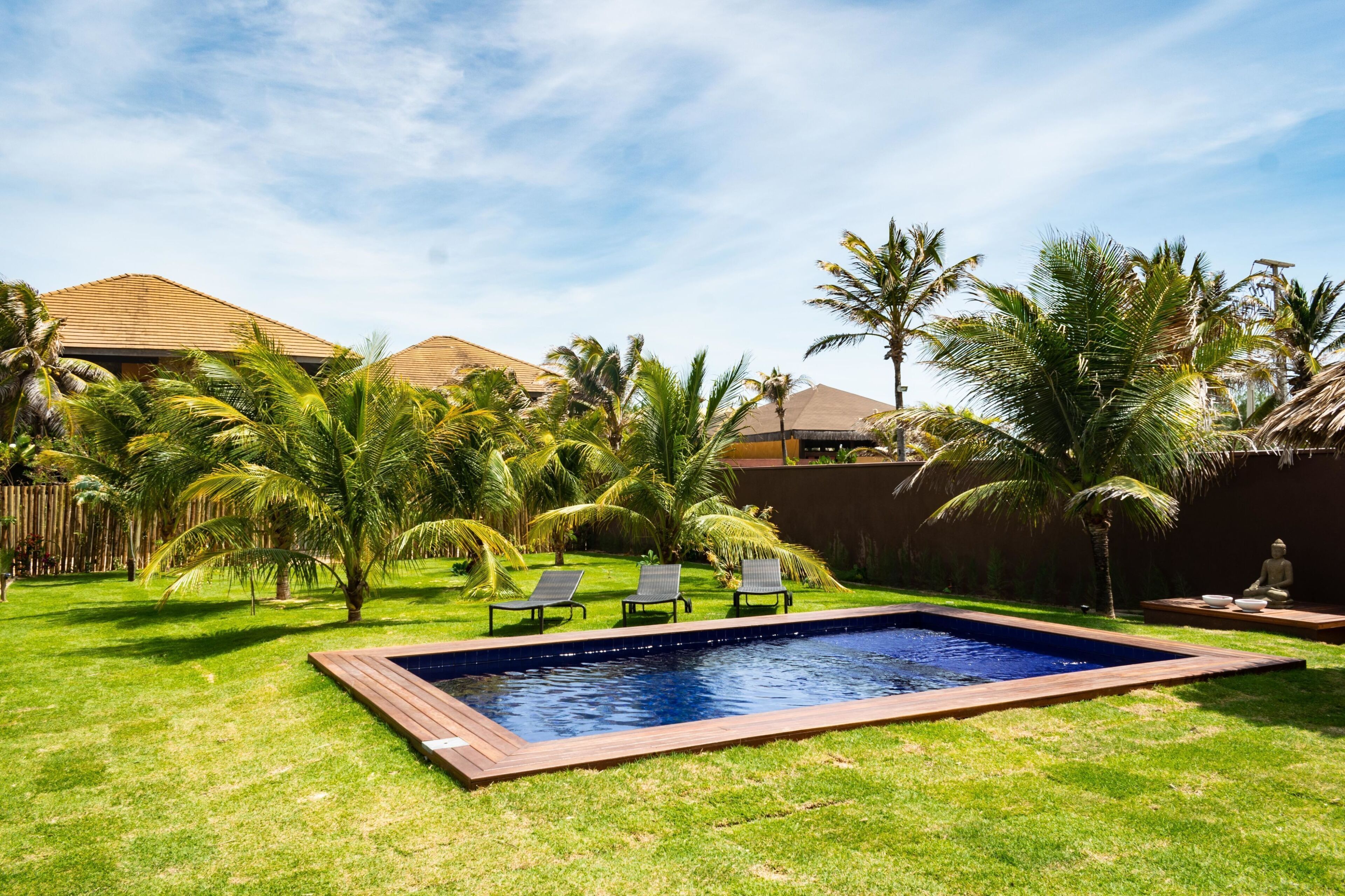 A swimming pool with a wooden deck, surrounded by palm trees and a grassy lawn.