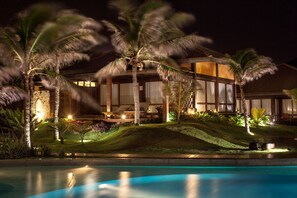 A nighttime view of a resort with a swimming pool, palm trees, and a well-lit building with large windows.