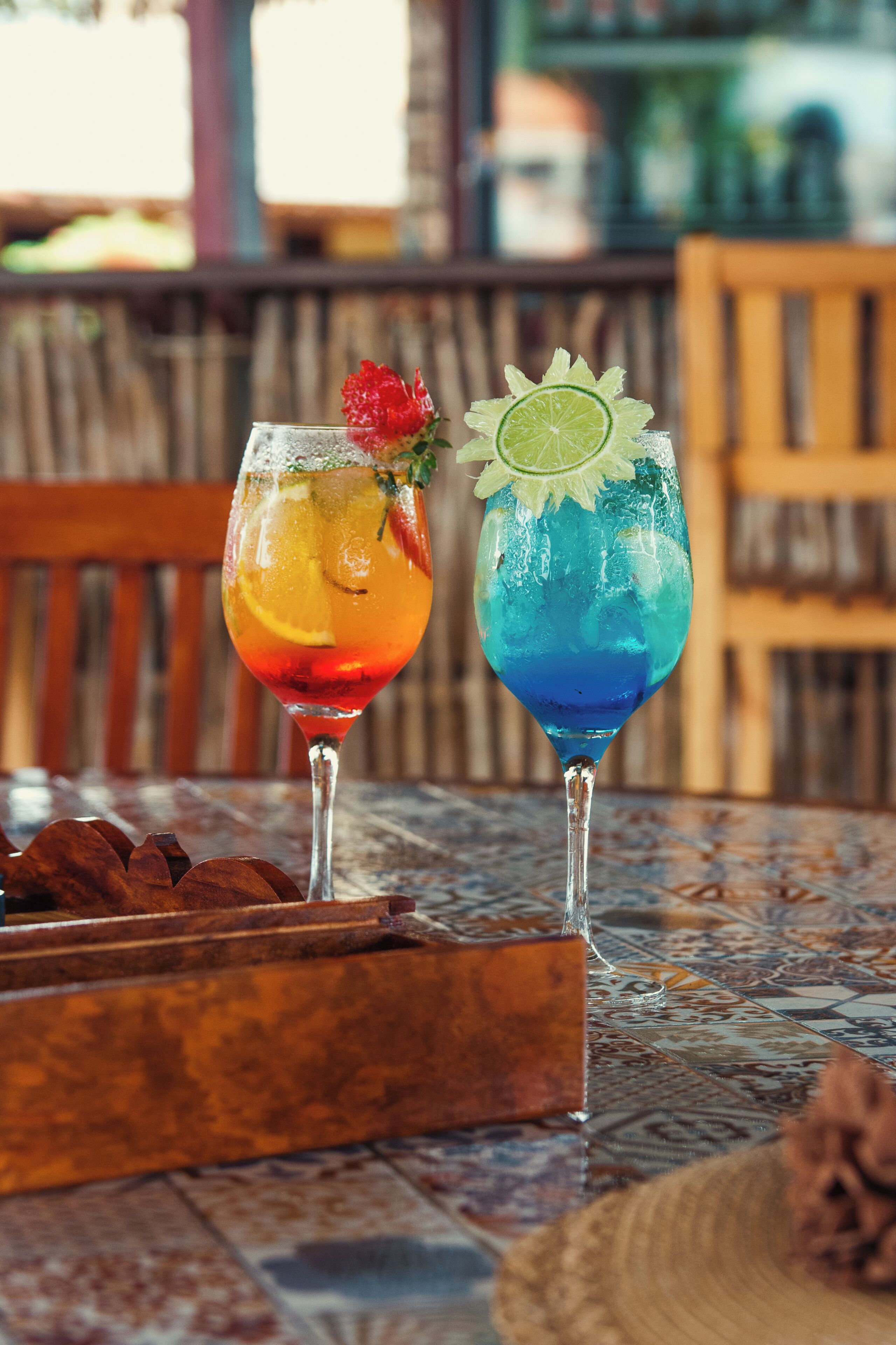 Two cocktails on a table with a patterned tablecloth, a wooden chair, and a wooden tray with what appears to be nuts.