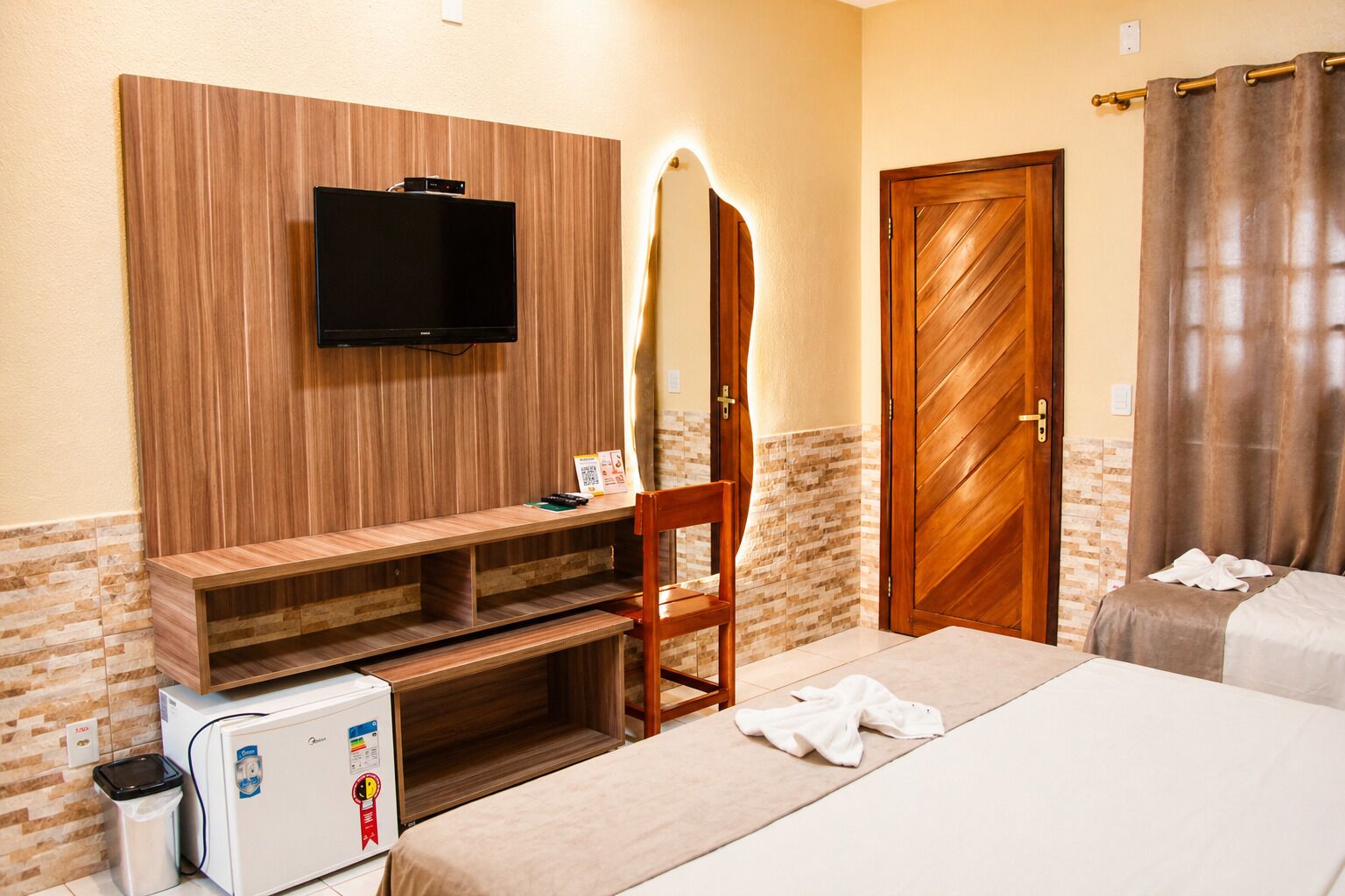Ein Hotelzimmer mit einem an der Wand befestigten Fernseher, einem Holztisch, einem Stuhl, einem Bett mit Handtüchern und einer Tür mit Spiegel.