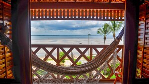 Una hamaca en un balcón con vista a una playa, palmeras y cielo nublado.