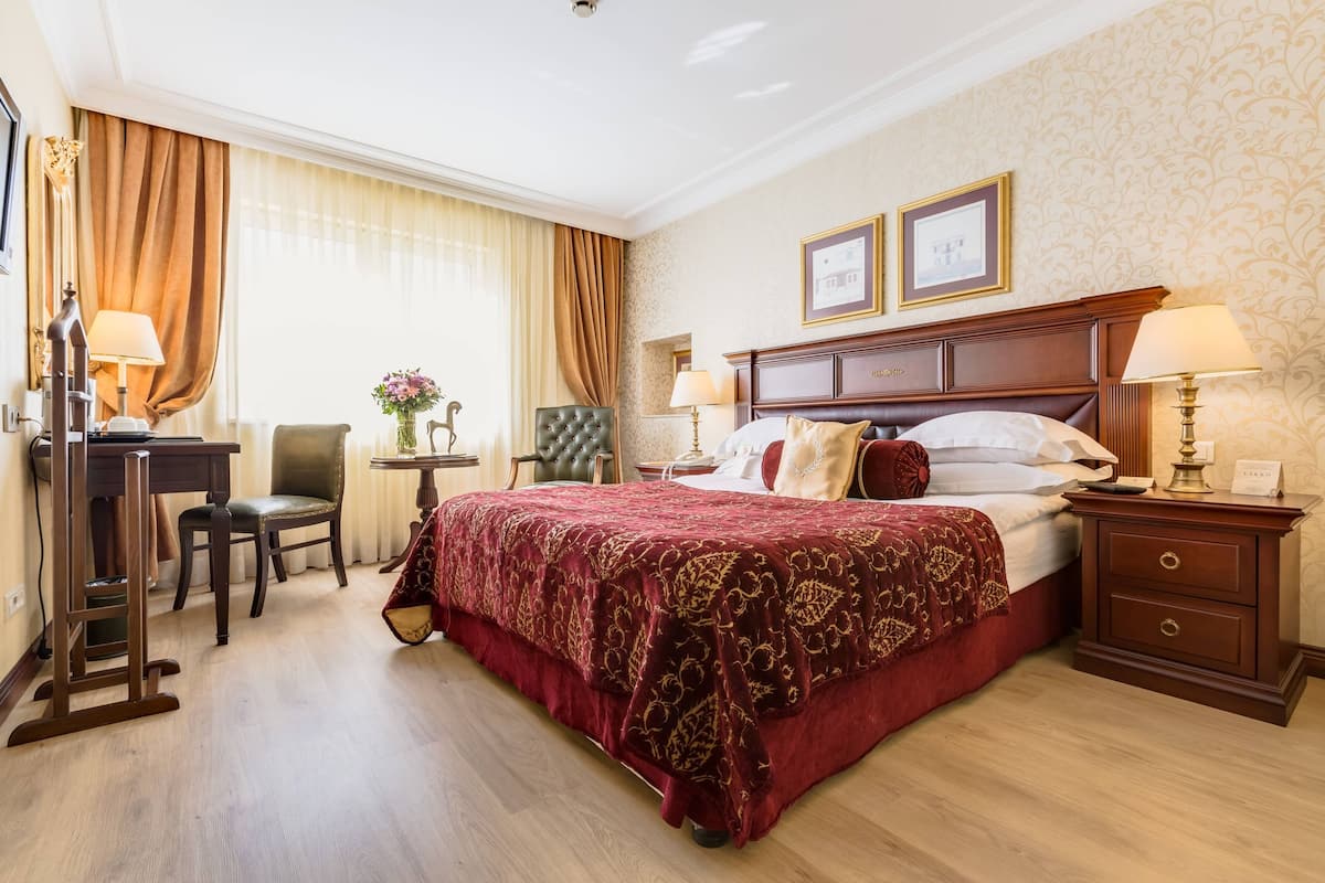 Una habitación de hotel con una cama grande, un escritorio, una silla, una mesita con un jarrón y dos cuadros en la pared.