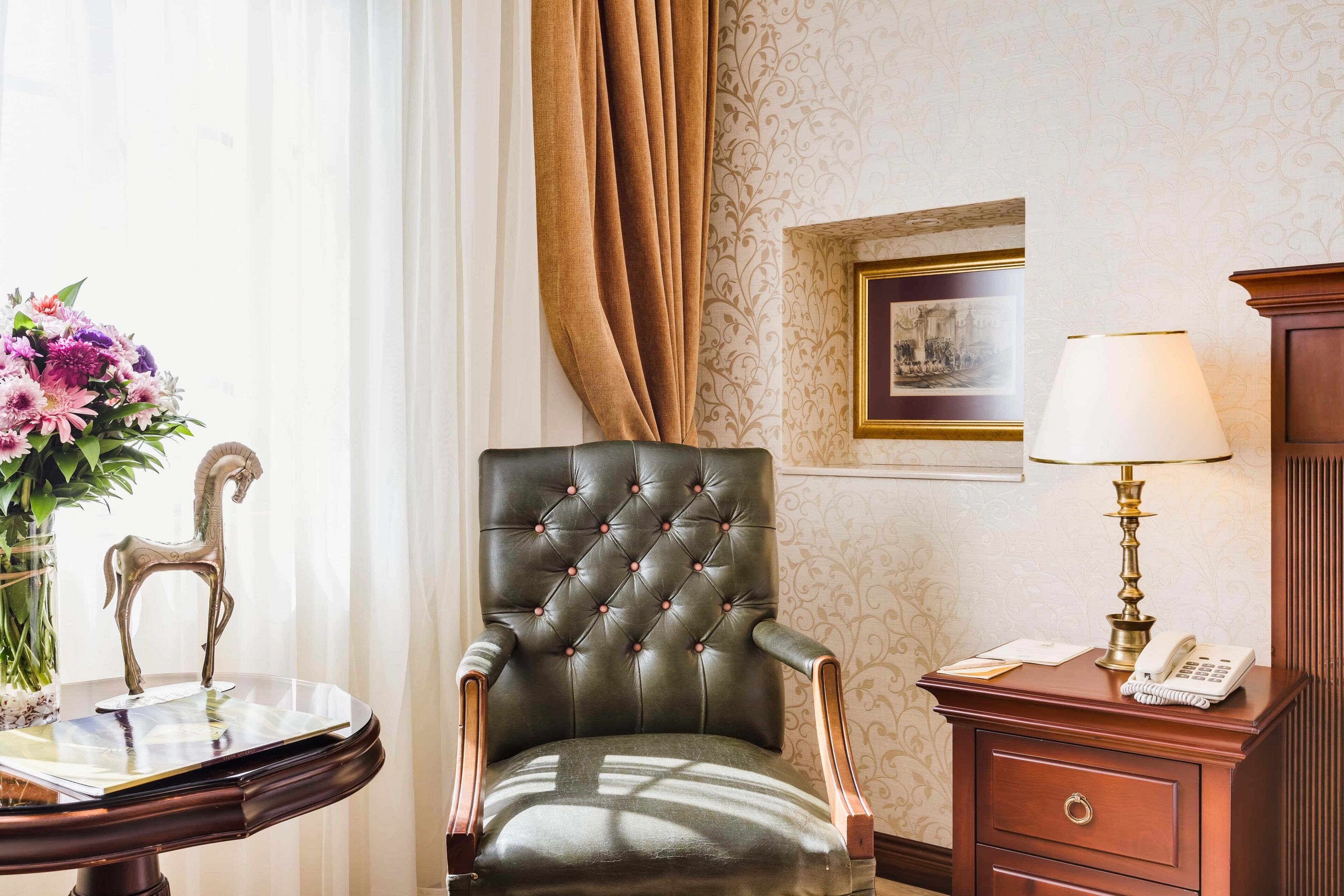 Une pièce comprenant un fauteuil en cuir capitonné, une petite table sur laquelle est posé un vase de fleurs, une lampe, un tableau encadré et une table de chevet en bois avec un téléphone.