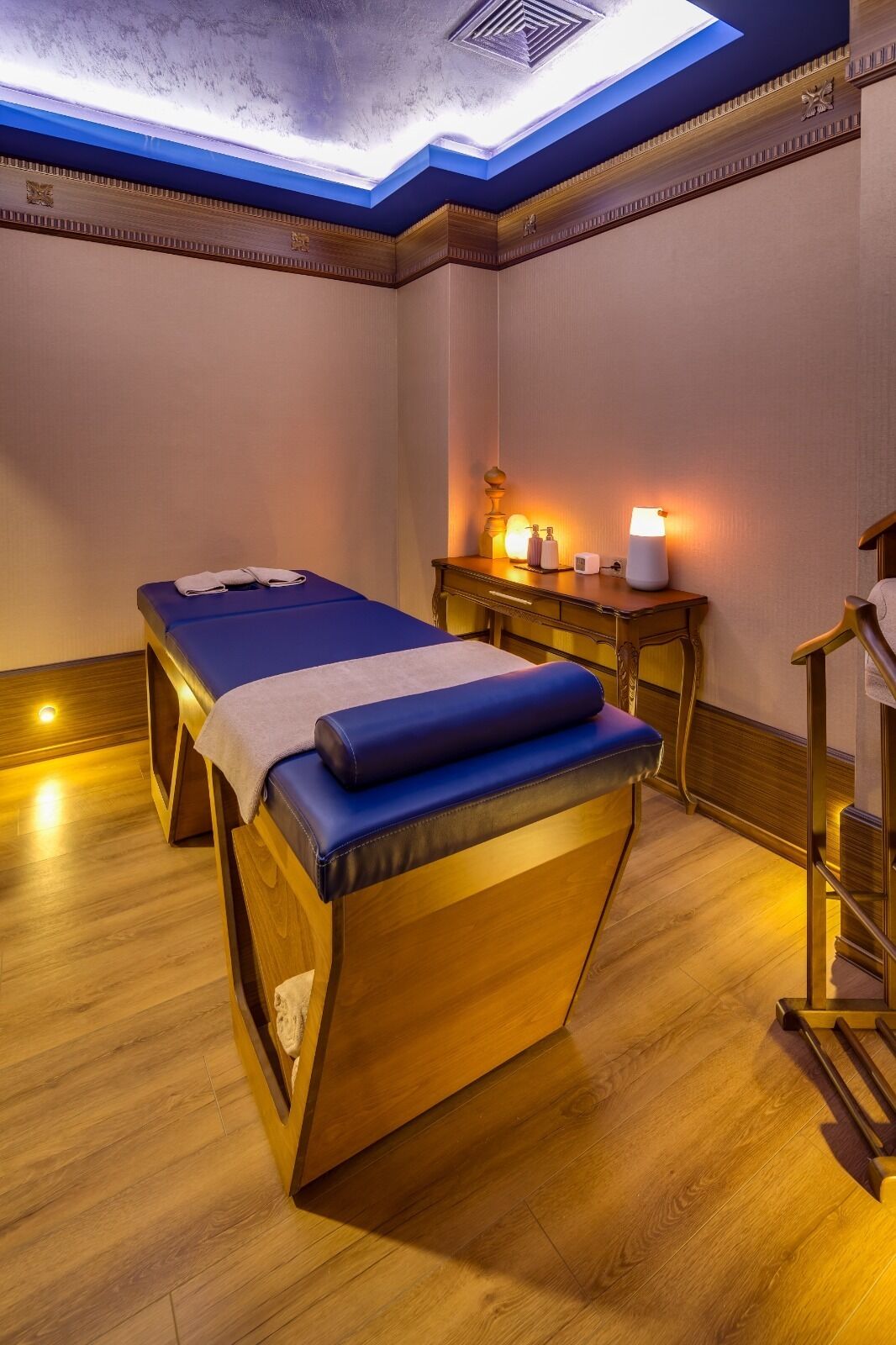 A room with two massage tables, a wooden bench, a small table with a lamp, and a wall with a decorative ceiling.