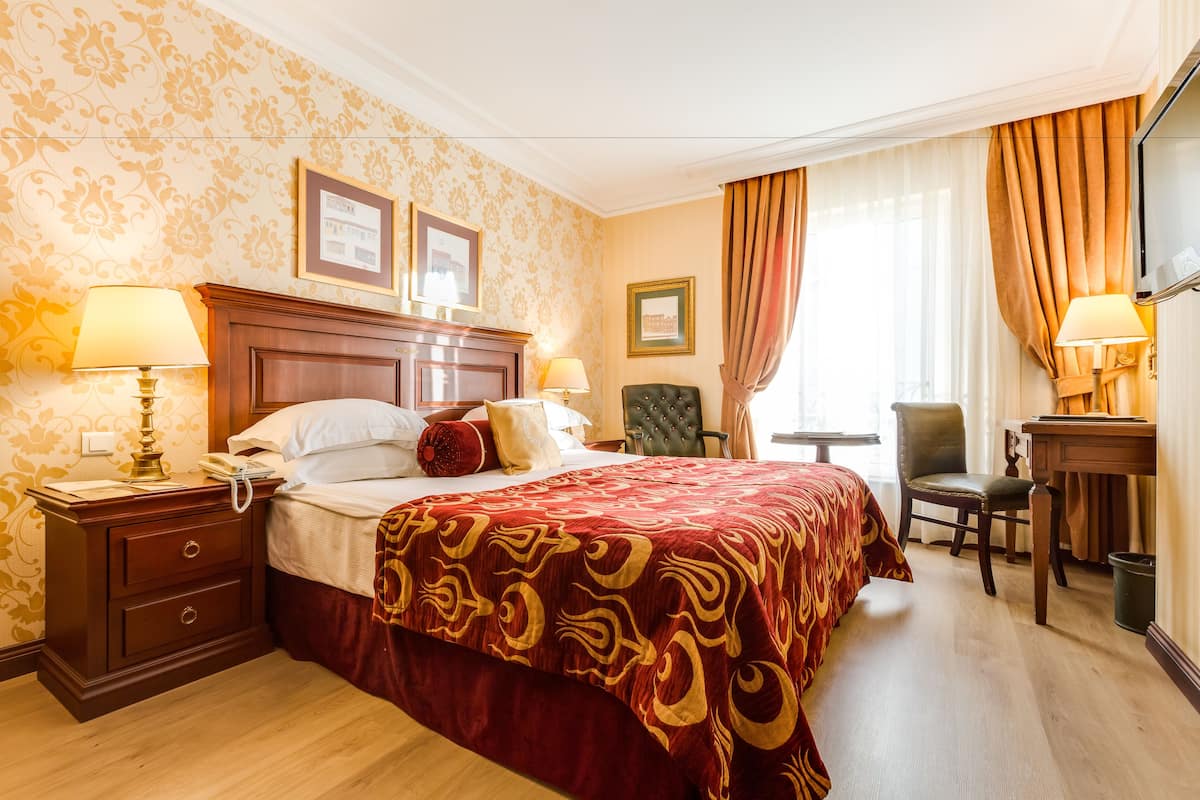 Habitación de hotel con una cama grande, dos mesitas de noche con lámparas, un escritorio pequeño, una silla y un ventanal con cortinas.