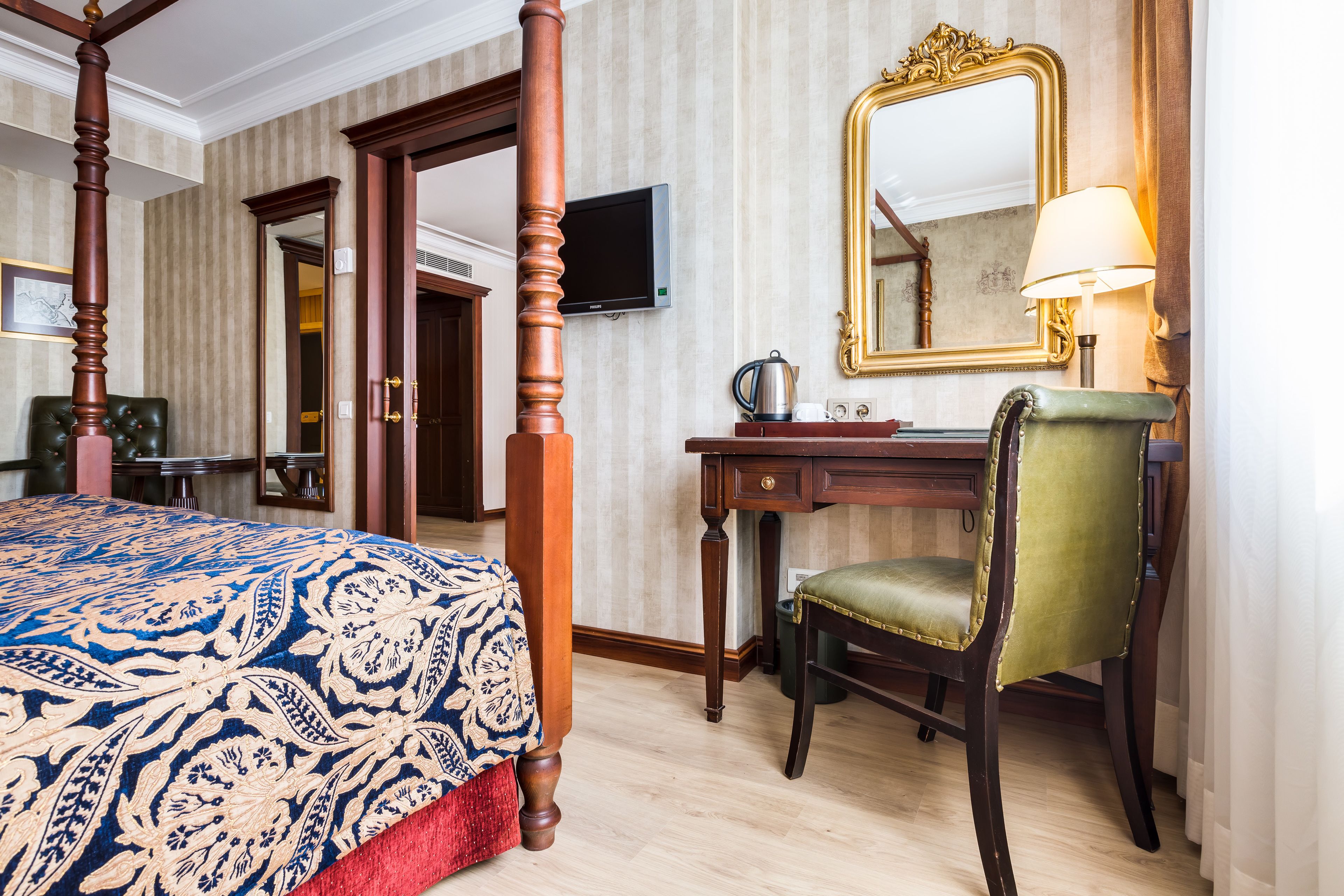 A four-poster bed with a patterned bedspread, a desk with a lamp, a green upholstered chair, and a large mirror on the wall.