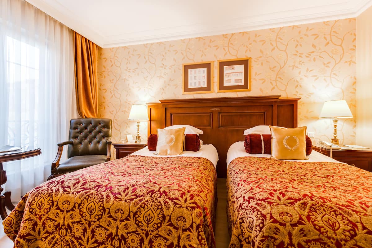 Habitación de hotel con dos camas, un cabecero de madera, un sillón de cuero y dos mesitas de noche con lámparas.