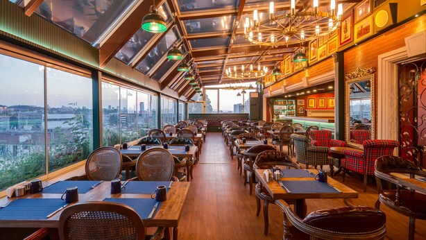 A restaurant with high ceilings, large windows, and a view of the city. Tables are arranged for dining, and there are chairs and tables for guests. - Gordion Hotel - Special Class (Ankara)