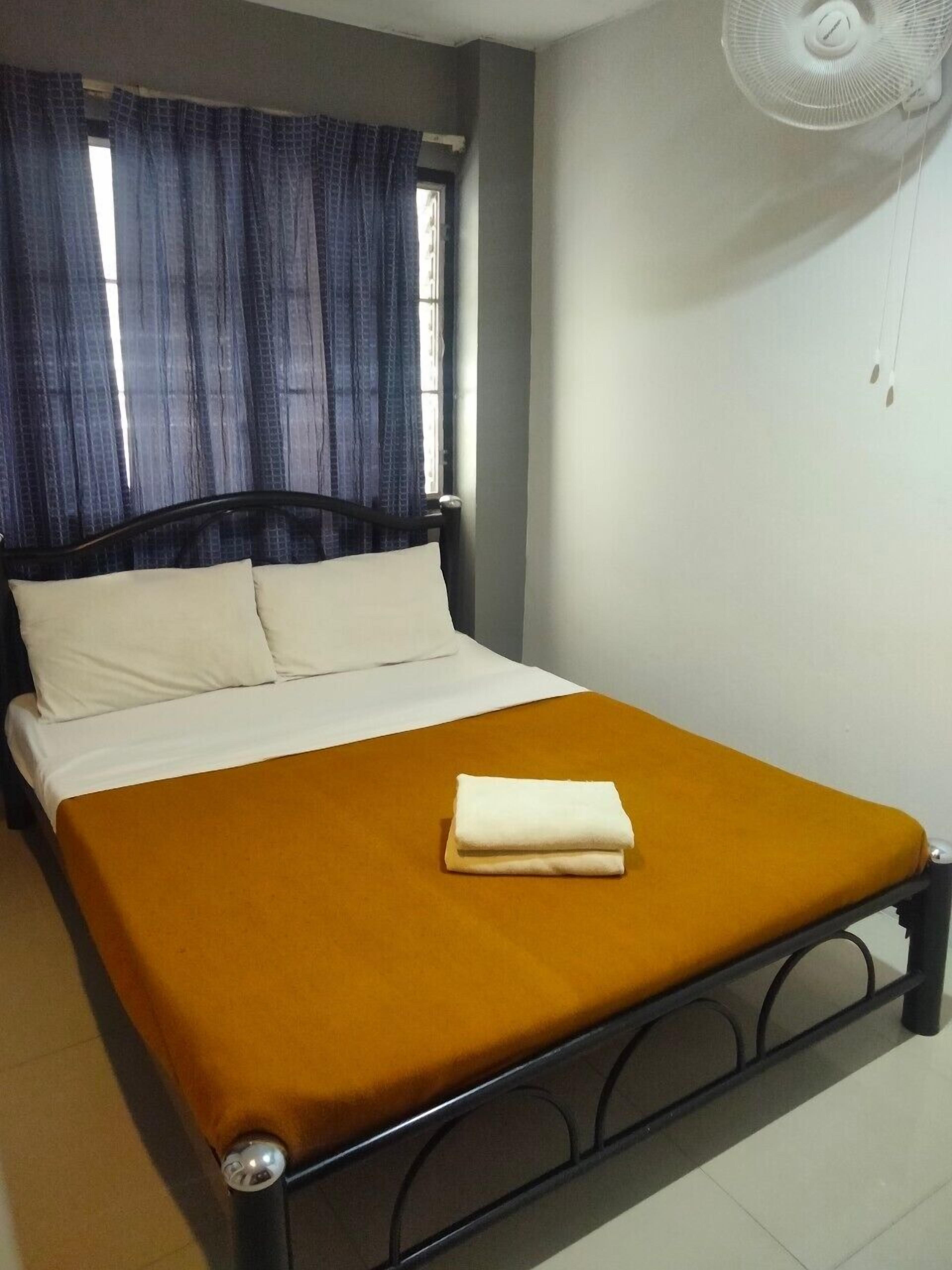 Un letto singolo con materasso bianco e coperta color senape, struttura in metallo nero, ventola a muro bianca e finestra con tende azzurre.