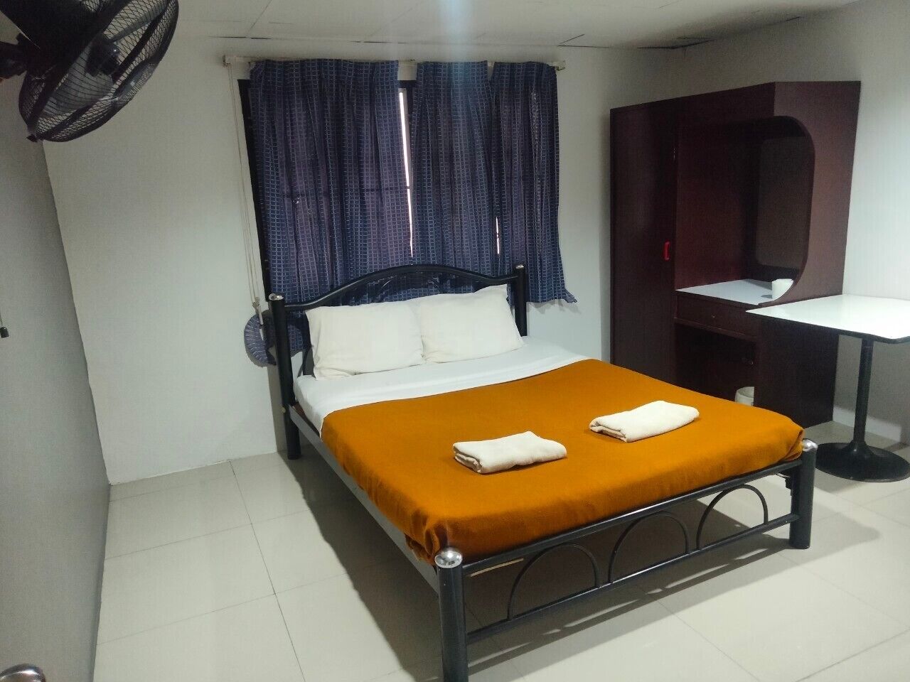 Una cama individual con colcha naranja y almohadas blancas, una mesita pequeña y un armario de color oscuro en una habitación con paredes blancas y suelo de baldosas.