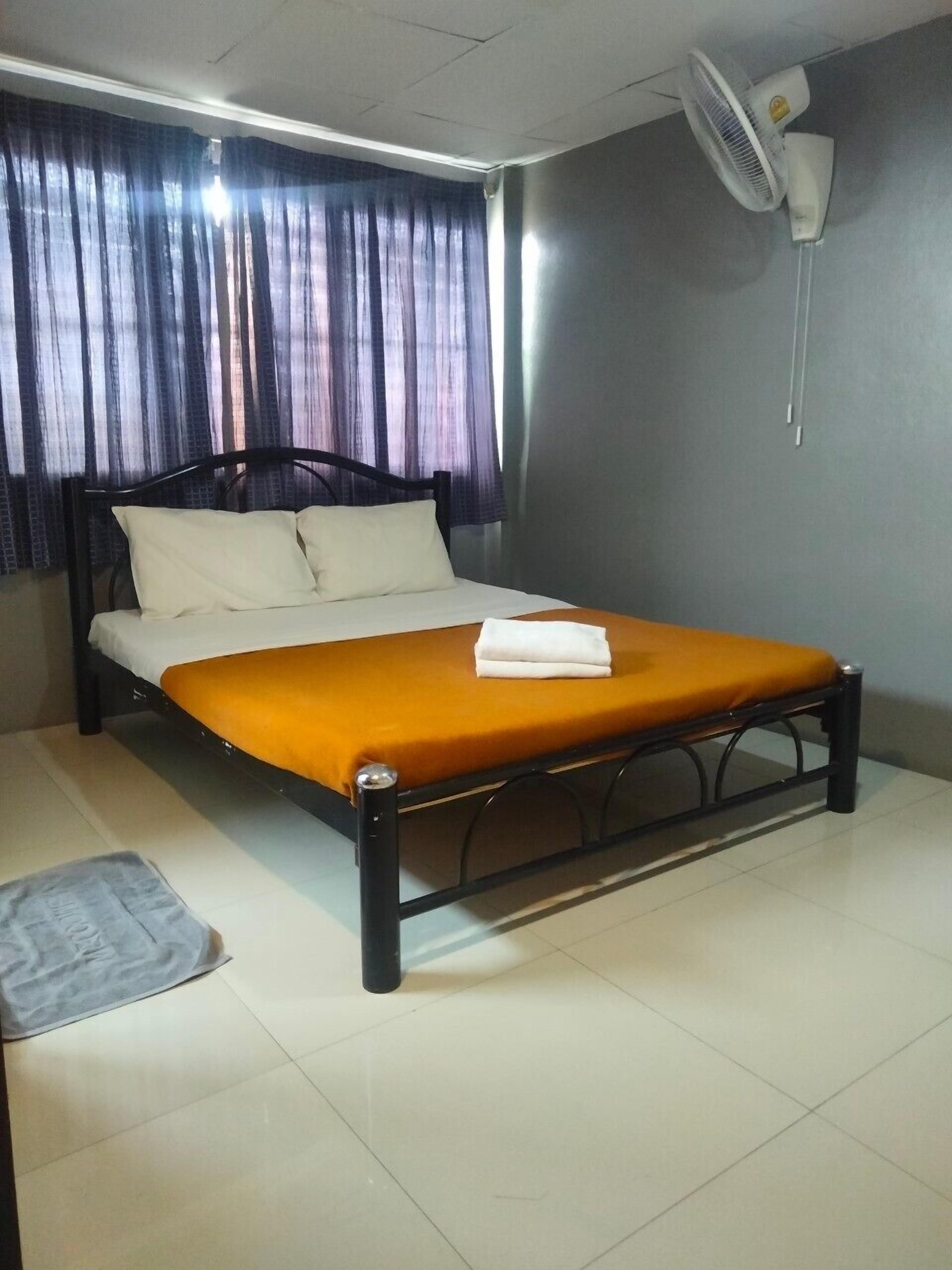 A single bed with a white mattress and an orange bedspread, a black headboard, a white wall fan, and a curtain partially drawn to the side.