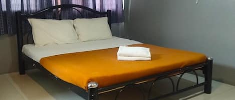Una cama individual con colchón blanco y colcha naranja, cabecera negra, ventilador de pared blanco y una cortina abierta a un lado.