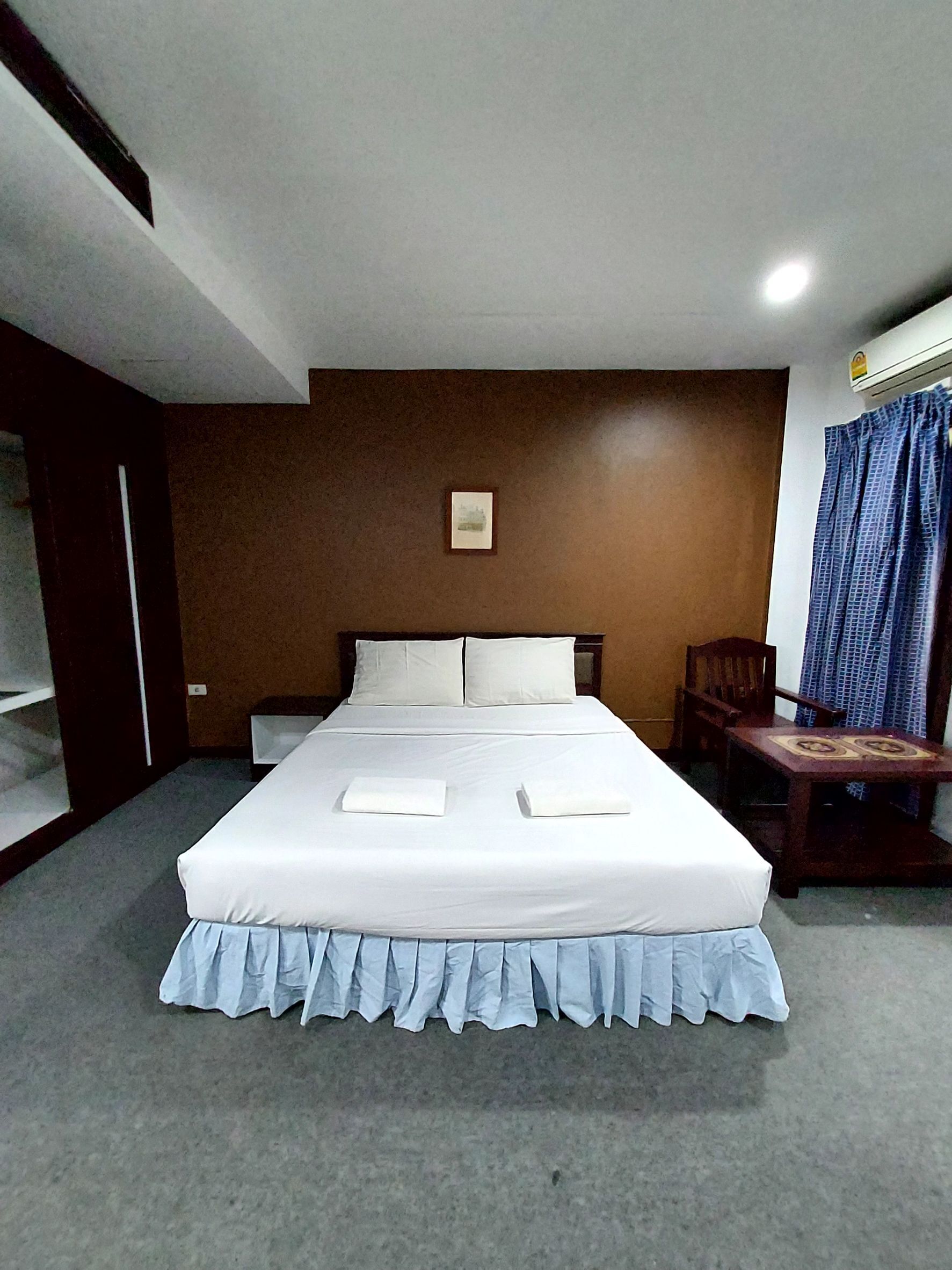 A neatly made bed with white linens and a blue skirted bed skirt. There is a wooden chair and a small table with a decorative top. A framed picture hangs on the wall above the bed.