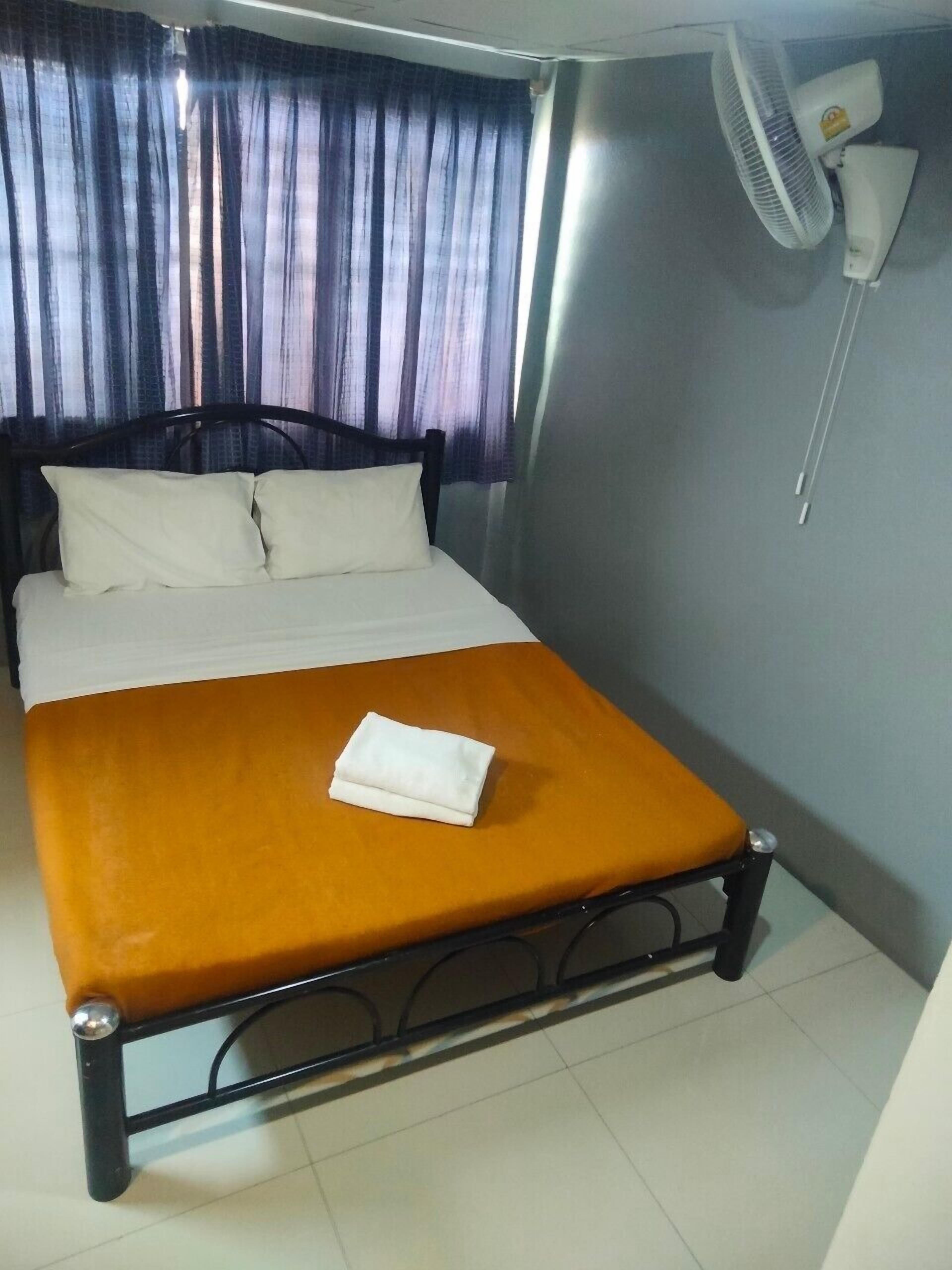A single bed with a white mattress and a folded white towel, a dark metal frame, a wall-mounted fan, and a window with purple curtains.