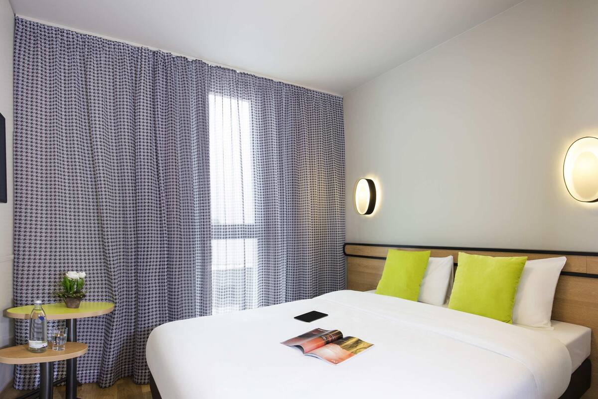 Een hotelkamer met een bed, nachtkastje, lamp en een raam met gordijnen.