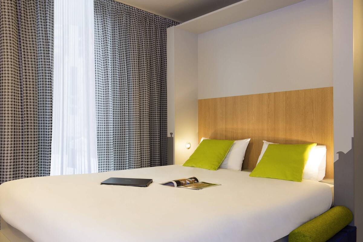 Een net opgemaakt bed met witte lakens en twee groene kussens, een laptop en een tijdschrift op het bed.
