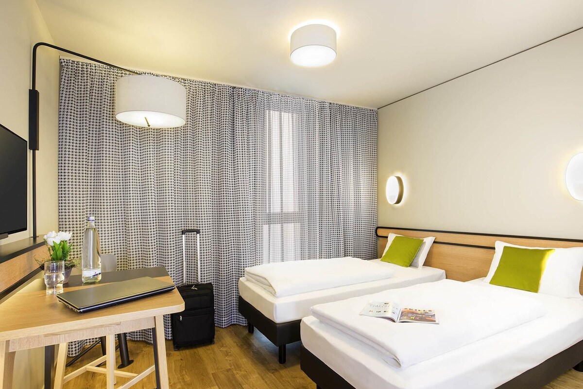 A hotel room with two beds, a desk, a chair, a TV, and a window with curtains.