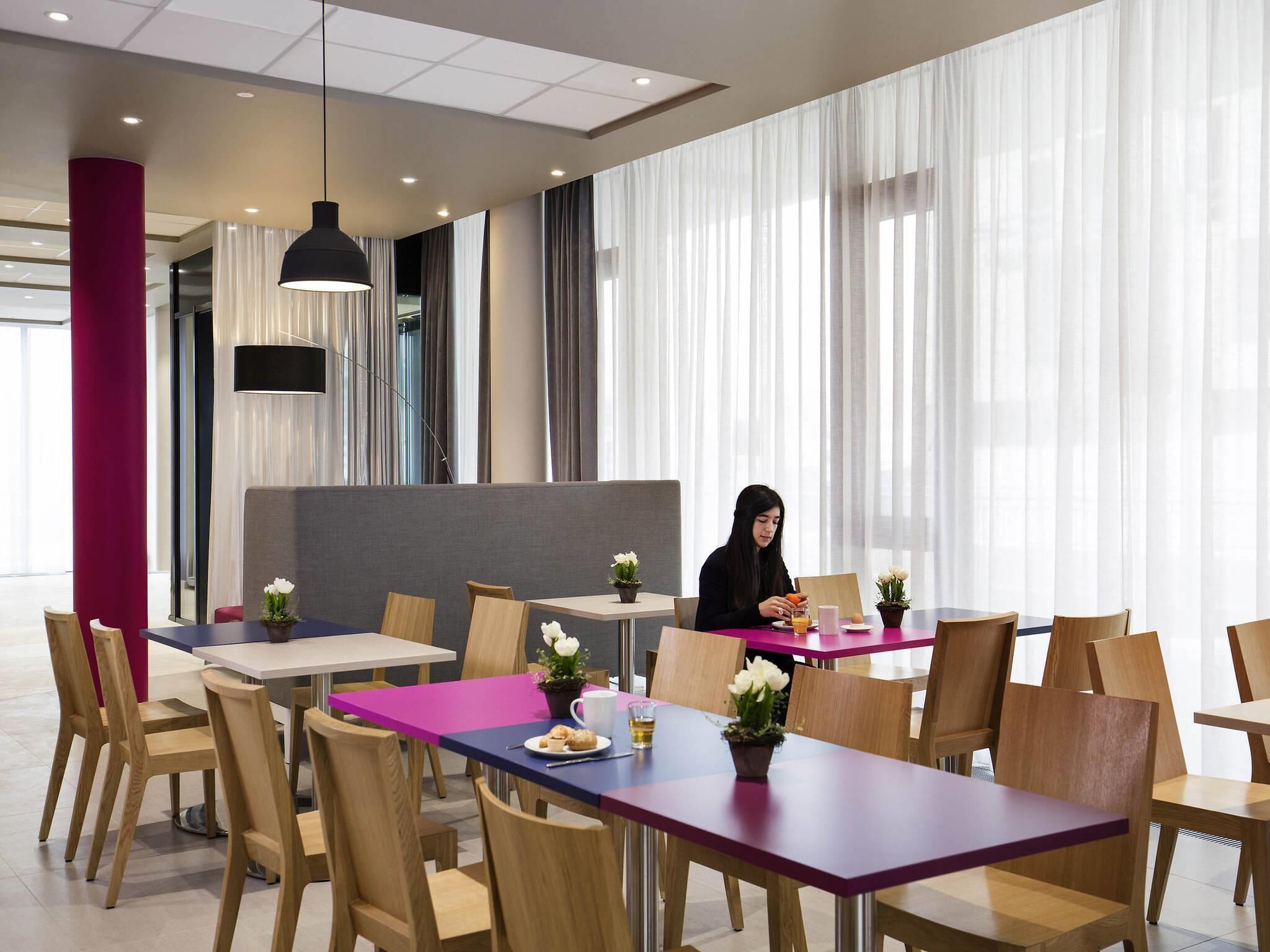 Un espace de restauration moderne, doté de plusieurs tables et chaises, d’une grande fenêtre ornée de rideaux légers et d’un pilier rouge.