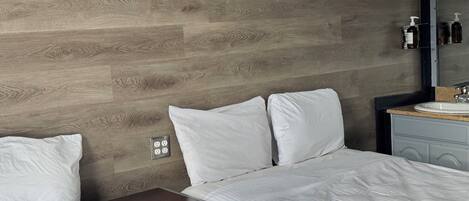 Habitación de hotel con pared de madera, cama con ropa blanca, mesita de noche, espejo y lavamanos.