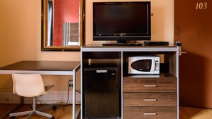 A compact hotel room with a desk, a flat-screen TV, a mini-fridge, a microwave, a white chair, and a door with the number 103. - Europa Hotel (San Francisco)