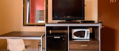 A compact hotel room with a desk, a flat-screen TV, a mini-fridge, a microwave, a white chair, and a door with the number 103.