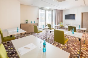 Een hotelvergaderruimte met witte tafels, groene stoelen en een grote televisie aan de muur.