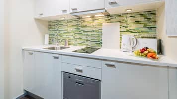 A modern kitchen with white cabinets, a stainless steel dishwasher, a fruit bowl with various fruits, and a green and white tiled backsplash.