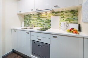 Una cuina moderna amb armaris blancs, un rentaplats d'acer inoxidable, un plat de fruita amb diferents fruites i una barra posterior amb rajoles verdes i blanques.