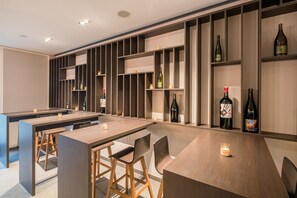 A modern bar area with wooden tables and chairs, shelves with bottles, and recessed ceiling lighting.