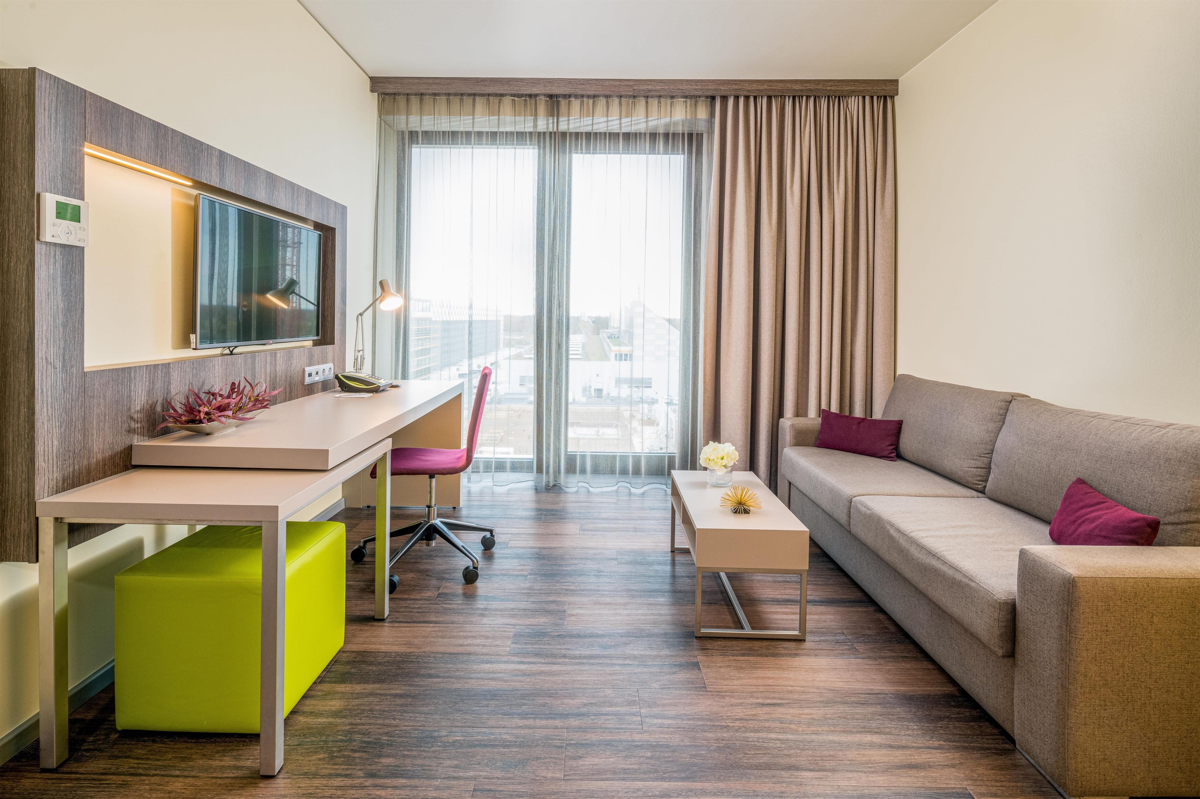 A hotel room with a desk, a chair, a sofa, a coffee table, and a large window with curtains.