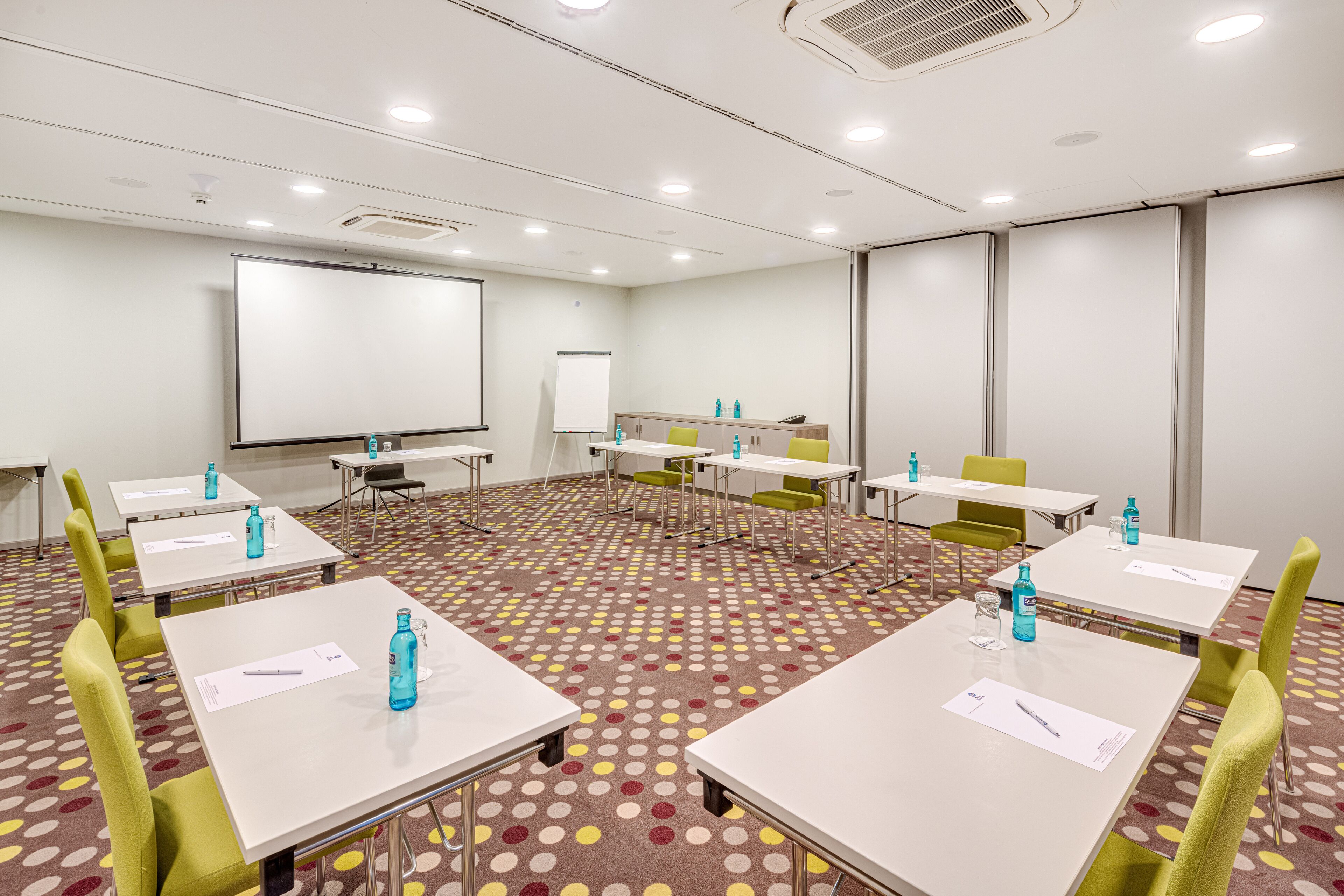 Ein Konferenzraum mit weißen Tischen, grünen Stühlen und einer großen Whiteboard-Fläche.