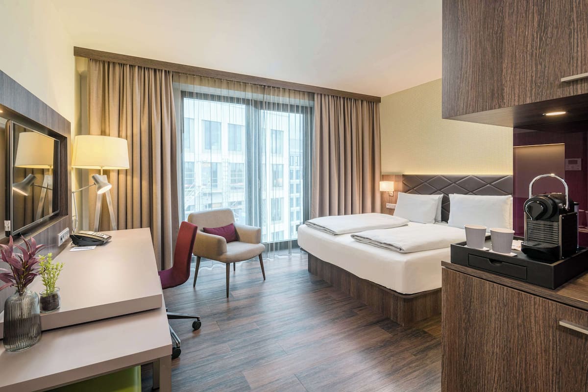 Habitació d'hotel moderna amb llit, escriptori, cadira i una finestra gran amb cortines.