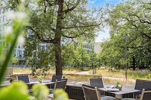 Een terras met tafels en stoelen, omgeven door bomen en een gebouw op de achtergrond.