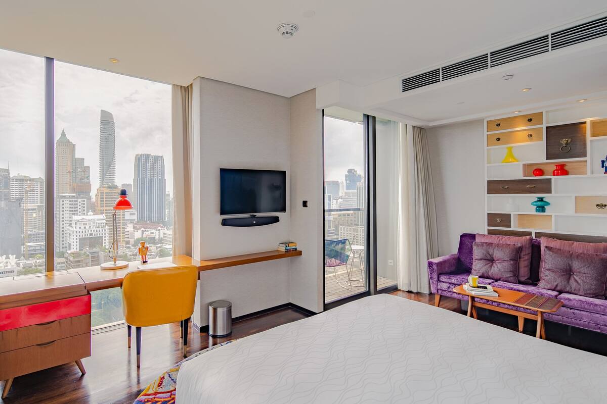 A modern hotel room with a large bed, a desk with a chair, a purple sofa, a TV mounted on the wall, and a city view through large windows.