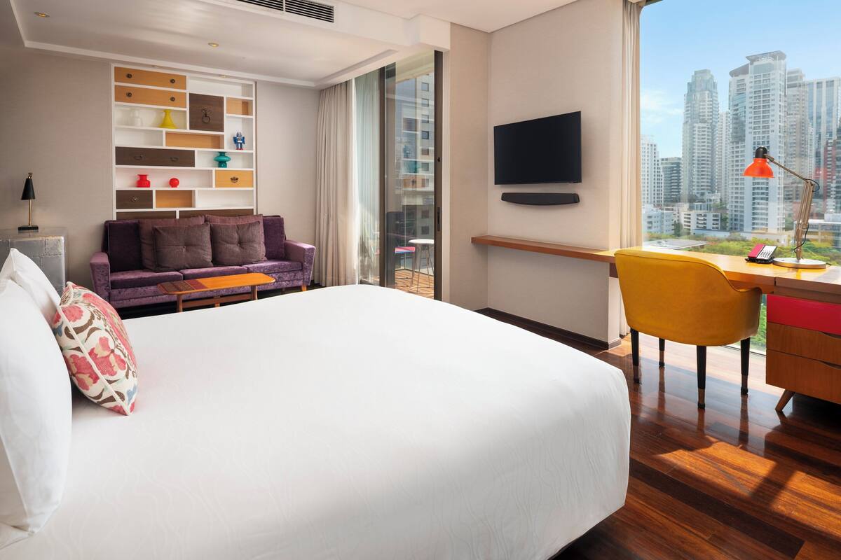 A modern hotel room with a large bed, a sofa, a desk with a chair, a TV, and a city view.