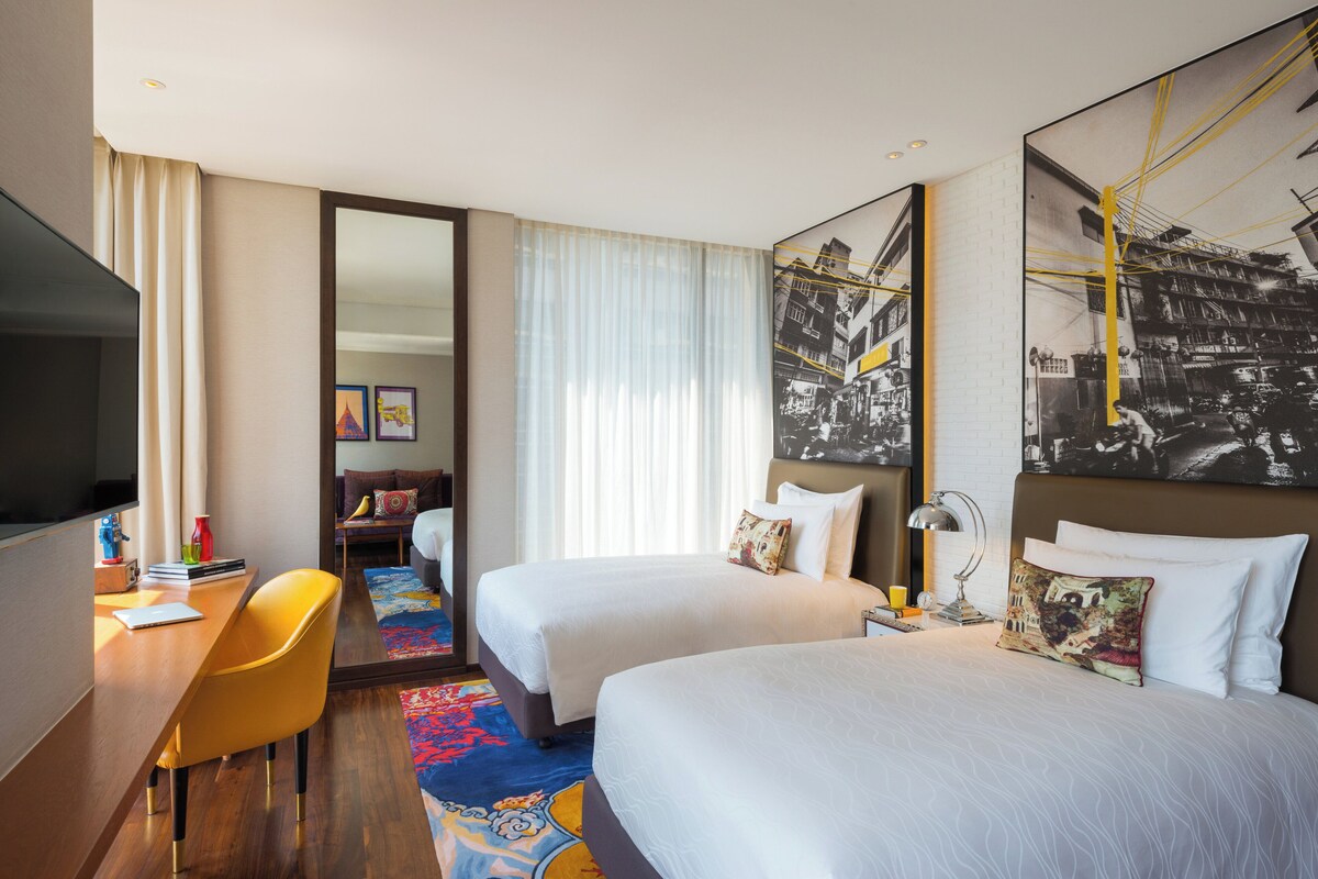 A hotel room with two beds, a desk, a chair, a TV, and a large mirror.