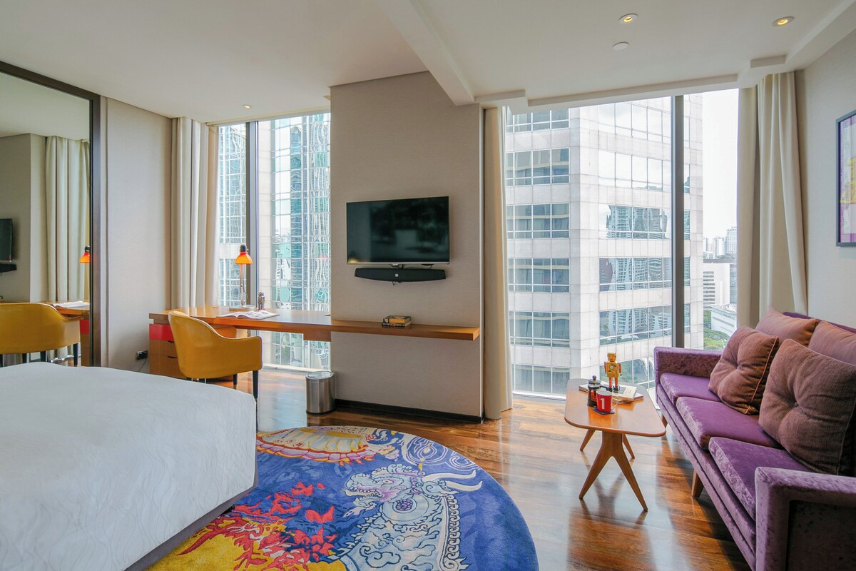 A hotel room with a bed, a desk, a sofa, a TV, and large windows with a city view.