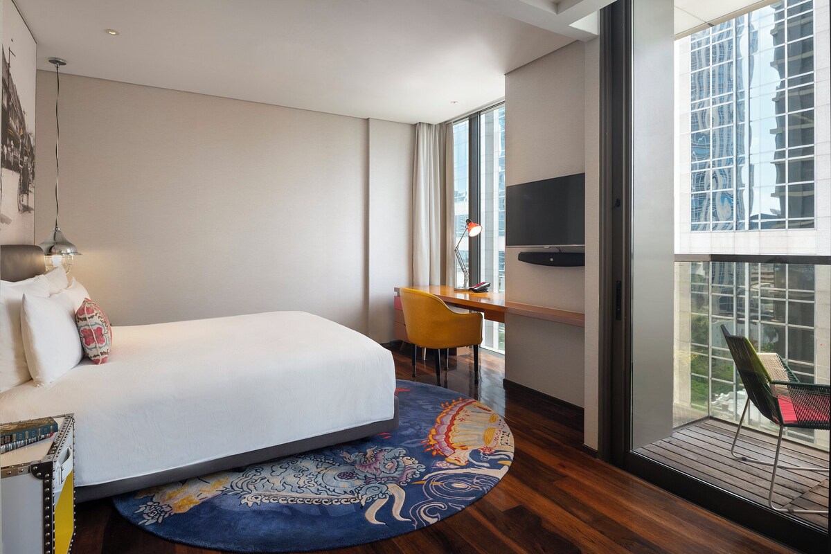 A modern hotel room with a large bed, a desk, a chair, and a balcony with a view of the city.
