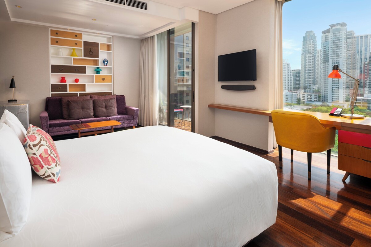 A modern hotel room with a large bed, a sofa, a desk with a chair, a TV, and a city view.