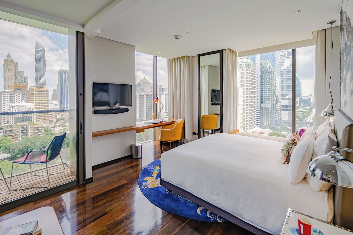 A hotel room with a large bed, a desk with a chair, a TV, and a balcony with a view of the city.